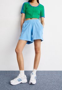 Green cropped zip-up top with blue accents, paired with light blue shorts, and white sneakers featuring blue stripes. Sock with scalloped edges.