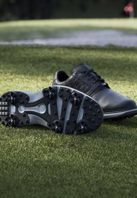 Chaussures de golf noires avec semelles à crampons sur de l'herbe verte près d'un piège de sable et un drapeau de trou de golf en arrière-plan.