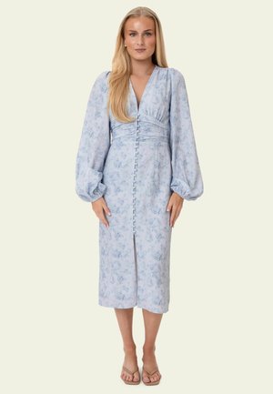 Kobieta o długich blond włosach, ubrana w jasnoniebieską sukienkę midi w kwiaty z bufiastymi długimi rękawami, guziczkami z przodu i rozcięciem, stojąca boso.