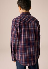 Rutig skjorta i marinblått och vinrött med vita linjer; har knäppta manschetter, krage och detalj på ryggens skoning. Tygets struktur ser slät ut.