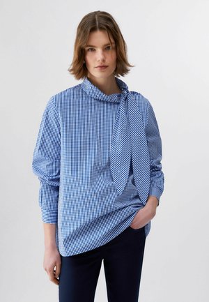 Mlajša ženska v modri in beli karo majici z detajlom kravate ter temnih hlačah, stoji pred enakomerno svetlim ozadjem.