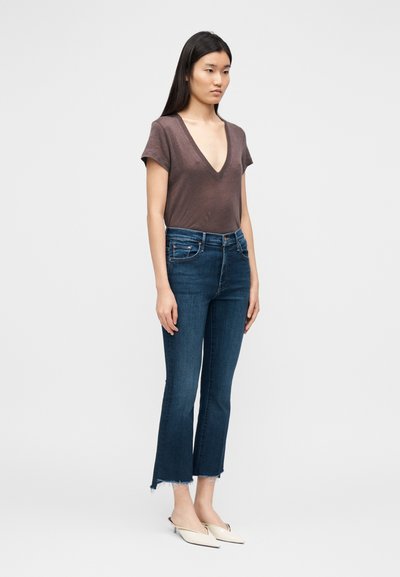 Tumesinised kõrge vöökohaga teksad narmendava äärega, kombineeritud vormi järgiva pruunika V-kaelusega särkiga. Modellil on jalas valged libisemisvabad mules.