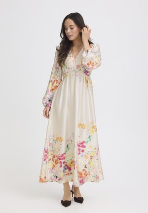 Žena si upravuje vlasy, oblečená v dlhých krémových kvetovaných šatách s nadýchanými rukávmi a čiernymi lodičkami, stojaca na bielom pozadí.