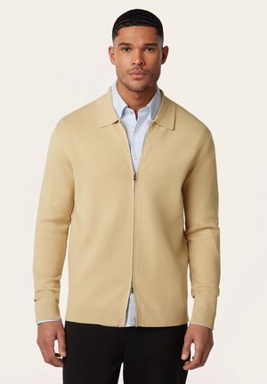 Man draagt een beige zipvest over een blauw overhemd met kraag, staand tegen een effen lichte achtergrond.