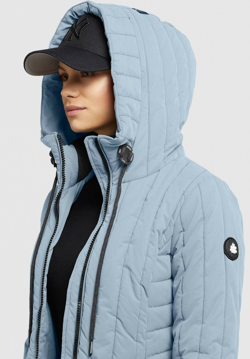 Hellblau gesteppte Jacke mit Kapuze; ausgestattet mit einem dunklen Reißverschluss, Zugbänder-Akzenten und einem Logo-Patch am linken Arm. Schwarze Mütze darunter getragen.