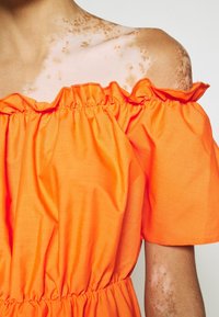 Personne portant une robe orange décolletée qui révèle la peau, avec des taches de rousseur brunes et des marques sur l'épaule et le bras.