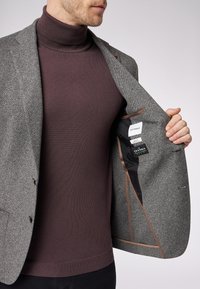 Gray herringbone blazer with brown interior lining, featuring two buttons and a sleek design. Worn over a dark mauve turtleneck sweater.