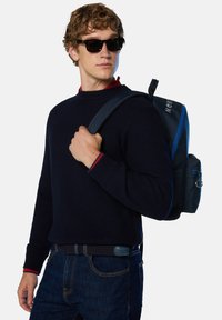 Suéter texturizado en azul marino, jeans oscuros y una mochila con acentos azules que tiene un bolsillo con cremallera, herrajes reflectantes y correas ajustables.
