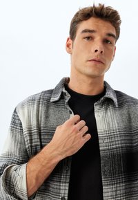 Camisa de cuadros en gris y blanco con cuello abotonado, que presenta una textura cepillada, llevada sobre una camiseta negra. Mano situada en el cuello.