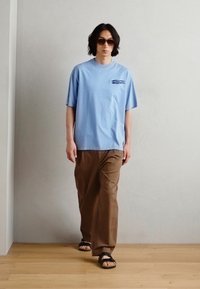 Tricou din bumbac de culoare albastru deschis, cu un buzunar pe piept, asortat cu pantaloni largi de culoare maronie și sandale negre. Textură netedă, design casual.