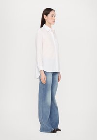 Witte langarmshirt met een subtiele glans en een borstzak, gecombineerd met wijde lichtblauwe denim jeans en zwarte schoenen.