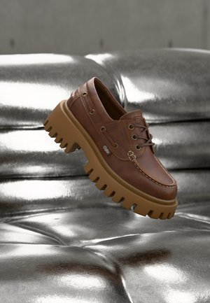 Chaussure marron en cuir à lacets avec une semelle épaisse et robuste de couleur beige, flottant au-dessus d'une surface en cuir argenté brillante et capitonnée.