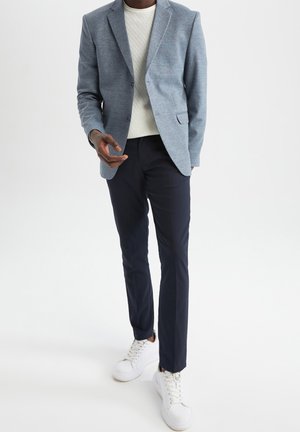 Mężczyzna ubrany w jasnoniebieską marynarkę, biały sweter dzianinowy, ciemnogranatowe spodnie i białe sneakersy, stojący na tle jednolitego białego tła.