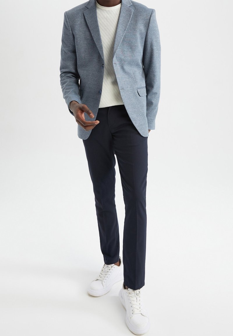 Homme portant un blazer bleu clair, un pull tricoté blanc, un pantalon bleu marine foncé et des baskets blanches, debout devant un fond blanc uni.