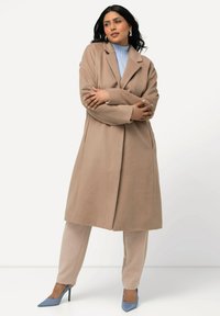 Cappotto di lana beige con vestibilità rilassata, collo a punte, maniche lunghe, abbinato a pantaloni grigi chiari e texture e scarpe con tacco blu.