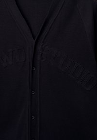 Schwarze Strickjacke mit V-Ausschnitt, ausgestattet mit strukturiertem "STUDIO"-Stickerei. Sie hat Knöpfe auf der Vorderseite und gerippte Kanten. Aus weichem Strickstoff.