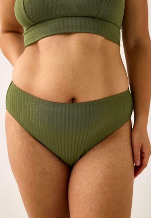 Close-up af en person iført olivengrønne, ribstrikkede bikinibunddele og en matchende crop top, der viser en afslappet holdning og naturlig hudtekstur.
