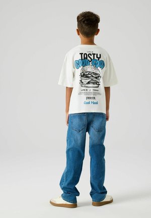 Chłopiec stojący tyłem do kamery, ubrany w białą koszulkę z nadrukiem burgera i niebieskim napisem, niebieskie dżinsy oraz białe trampki.