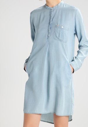 Lichtblauwe denimjurk met lange mouwen, mandarijnkraag, een borstzak aan de voorkant en zijzakken, gedragen met handen in de zakken.
