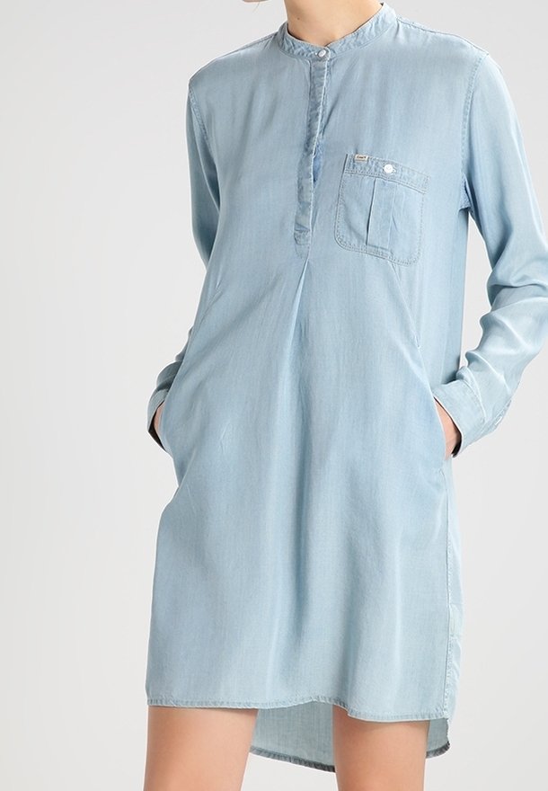 Robe en denim bleu clair à manches longues avec col mandarin, poche poitrine avant et poches latérales, portée avec les mains dans les poches.
