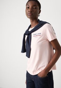 Chemise rose à manches courtes portant le texte "GANT 1949" en marine, accompagnée d'un pull marine drapé autour des épaules, assortie à un jean foncé.