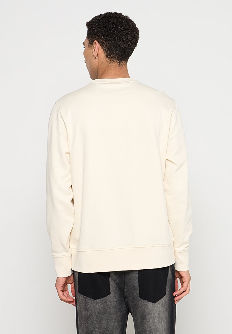 Cremefarbener Sweatshirt mit rundem Ausschnitt, gerippten Bündchen und Saum. Hat eine glatte Textur und wird über dunklen, abgetragenen Jeans getragen.