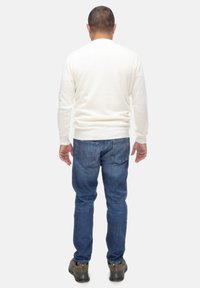 Maglione bianco a maglia con scollo rotondo, abbinato a jeans in denim blu. La persona indossa calzature marroni, vista da dietro su uno sfondo chiaro.