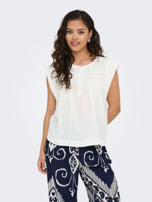 Jeune femme aux longs cheveux bouclés portant un chemisier blanc sans manches et un pantalon large bleu marine à motifs, debout devant un fond uni.