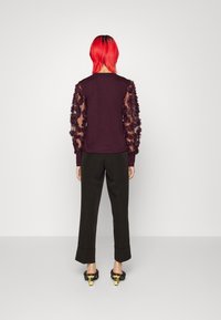 Persona con capelli rosso brillante, rivolta di spalle, indossa una maglietta viola con maniche floreali trasparenti, pantaloni neri con risvolto e tacchi neri con tacco dorato.