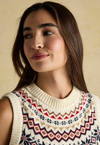 Adīta bezpaliku džemperis ar krēmīgu bāzi un daudzkrāsainiem ģeometriskiem rakstiem sarkanā, zilā, brūnā un tumši zilā krāsā ap kakla izgriezumu.