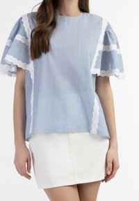 Una donna indossa un top azzurro chiaro con maniche a sbuffo bordate in pizzo e una gonna bianca, mostrando un look rilassato e alla moda.