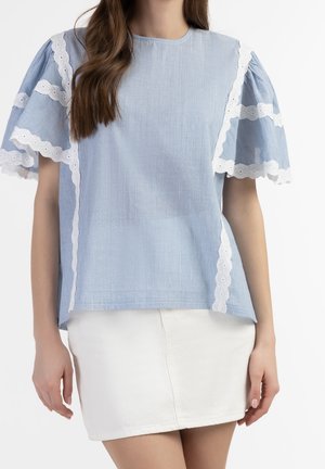 A woman wears a light blue top with lace-trimmed, puff sleeves and a white skirt, showcasing a relaxed, stylish look.