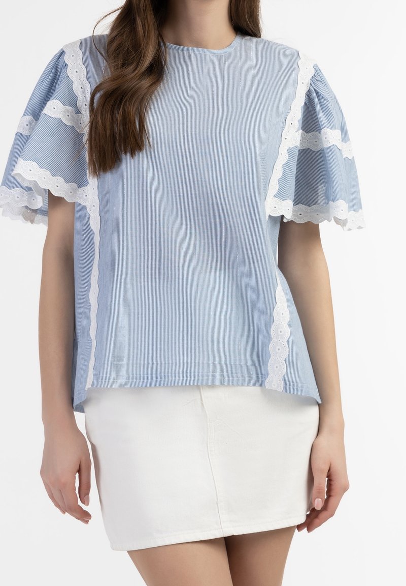 Una donna indossa un top azzurro chiaro con maniche a sbuffo bordate in pizzo e una gonna bianca, mostrando un look rilassato e alla moda.