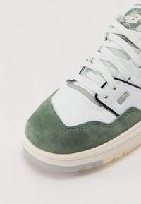 Baltos sportbačiai su žalia suede akcentu, tekstūruotu tinkleliu ir lygiaverčiais odiniais elementais. Yra juodų detalių ir pilkos guminės pado.