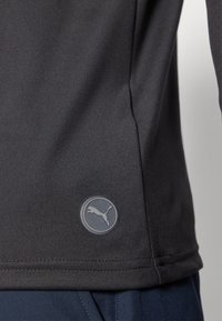 Schwarzes Sporthemd mit glatter Textur, versehen mit einem runden grauen Logo an der unteren Seite. Nahtdetails am Saum sind sichtbar.