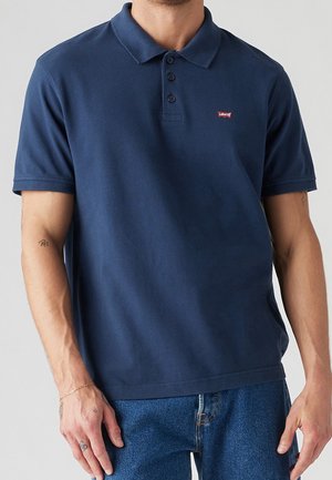 Mand iført en mørkeblå Levi’s polo med tre knapper og korte ærmer, kombineret med blå jeans.