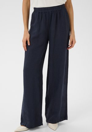 Femme portant un pantalon large bleu marine foncé avec une taille élastique et des chaussures blanches pointues, debout devant un fond uni.
