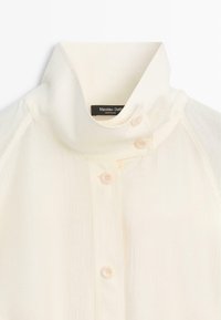 Crèmekleurige blouse met hoge hals en gestructureerde stof, voorzien van vier crèmekleurige knopen aan de voorkant en een labeltje aan de kraag.