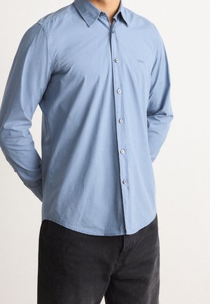 Homme portant une chemise bleu clair à boutons avec col et manches longues, associée à un pantalon noir, debout les mains dans le dos.