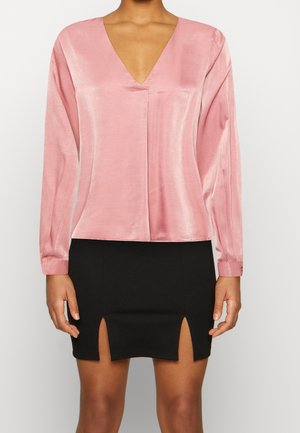 Frau trägt eine glänzende pinkfarbene Langarmbluse mit V-Ausschnitt und einen schwarzen Minirock mit Frontschlitzen, dargestellt von den Schultern bis zur Mitte des Oberschenkels.