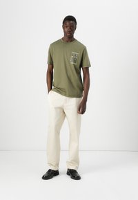 Un jeune homme se tient contre un fond blanc uni, portant un t-shirt vert olive avec un design de poche et un pantalon couleur crème.