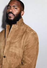 Tan ruskindsjakke med høj krave og minimal hardware. Har en glat tekstur og en enkelt knaplukning foran.