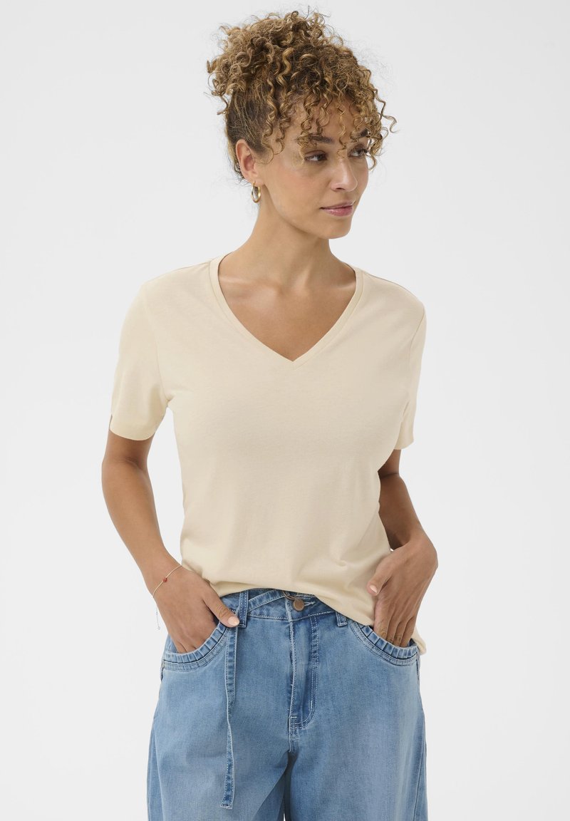 T-shirt a maniche corte con scollo a V in tessuto beige chiaro, caratterizzata da una vestibilità comoda e una texture liscia. Abbinata a jeans azzurri.