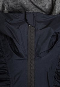 Námořnická bunda na zip s nařasenými detaily po stranách. Vyrobena z hladkého, lehkého materiálu; má vysoký límec a strukturovanou vnitřní látku.