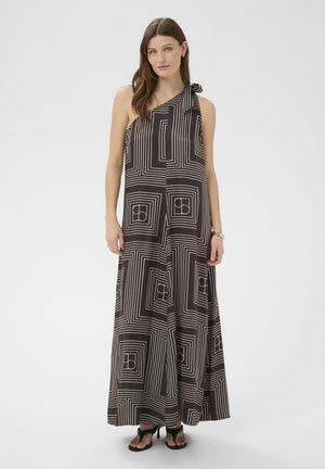 Kobieta w czarno-białej geometrycznej, asymetrycznej sukience maxi z wiązaniem i czarnych sandałach na obcasie, stojąca na jednolitym tle.
