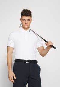 Camisa polo branca com colarinho e três botões, combinada com calças escuras e um cinto de fibra negra. Taco de golfe apoiado sobre o ombro.