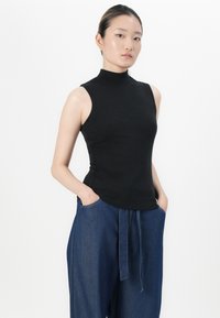 Mulher vestindo uma blusa preta de gola alta sem mangas e calças de ganga azuis largas, com as mãos nos bolsos, em pé contra um fundo branco.