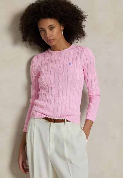 Frau mit lockigem Haar, die einen pinken Zopfpullover und weiße Hose mit einem geflochtenen braunen Gürtel trägt, steht vor einem schlichten Hintergrund.