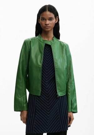 Jonge vrouw met een groene leren jas over een zwarte jurk met witte chevron- en verticale strepen, staand tegen een witte achtergrond.