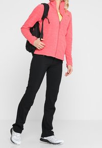 Veste en polaire rose zippée sur un haut jaune, pantalon noir et chaussures de sport blanches ; portant un sac à dos noir. Tenue décontractée pour l'extérieur.